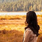 Woman in autumn landscape looking at serene lake, surrounded by colorful fall foliage.